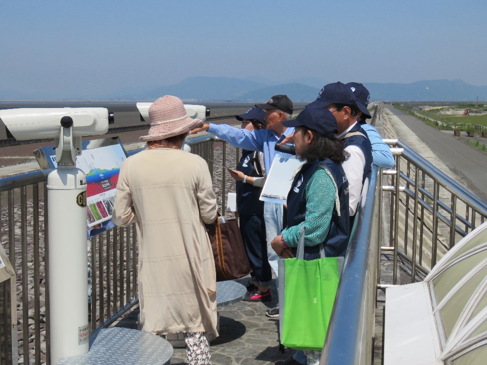晴天の下、展望台に設置された白い観光用望遠鏡の横で、解説パネルを囲みながらガイドの説明を熱心に聞く観光客たちの様子と、その背後に広がる広大な干潟や遠くの山々を、手前から奥にかけて構図に収めた写真