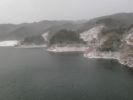 薄っすらと雪化粧を施された山々に囲まれた静かな湖畔の風景で、どんよりとした曇り空の下、冷たい水面が広がり、岸辺の木々や斜面が白く染まった冬の厳かで寂寥感のある自然の様子を捉えた写真
