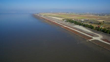 青々とした広大な海と干潟が左側に広がり、右側には海岸線に沿って真っ直ぐに伸びる堤防と、その先に広がる穏やかな田園風景や集落を、雲一つない青空の下で遥か上空から俯瞰で捉えた、開放感あふれる風景写真