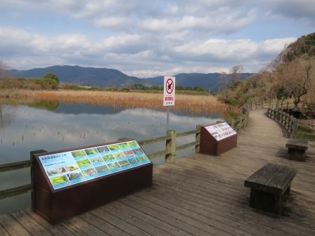 立て看板や案内板の奥に、池や茶色い枯れた植物が広がり、背景には山々がうつる写真