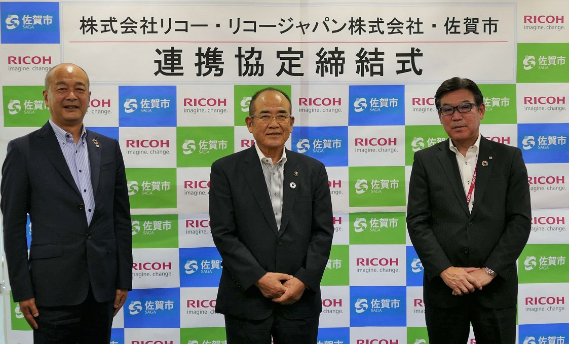 株式会社リコー、リコー・ジャパン株式会社、および佐賀市の三者が、連携協定の締結式で、企業ロゴと佐賀市のシンボルが並ぶパネルの前で並んで立っている写真