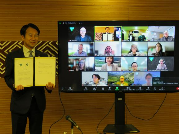 オンライン会議の様子が映し出された大型モニターの横で、スーツ姿の市長が署名済みの書面を広げて持ちながら、笑顔で正面を向いて立っている、国際的な提携や合意締結の場面を捉えた写真
