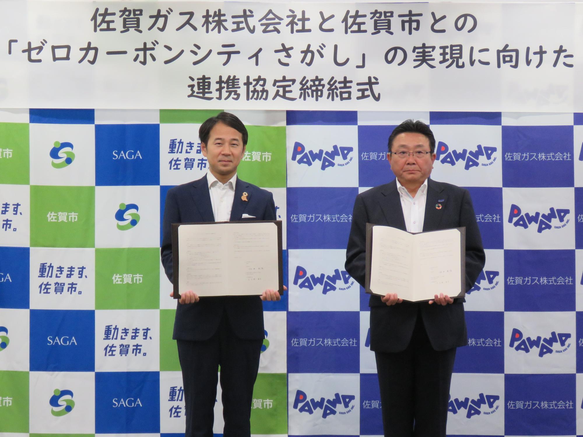佐賀ガス株式会社と佐賀市とのゼロカーボンシティさがしの実現に向けた連携協定締結式の横断幕の下、佐賀市と佐賀ガス株式会社のロゴが並ぶバックボードの前で、市長と大久保社長が協定書を掲げて記念撮影をしている写真