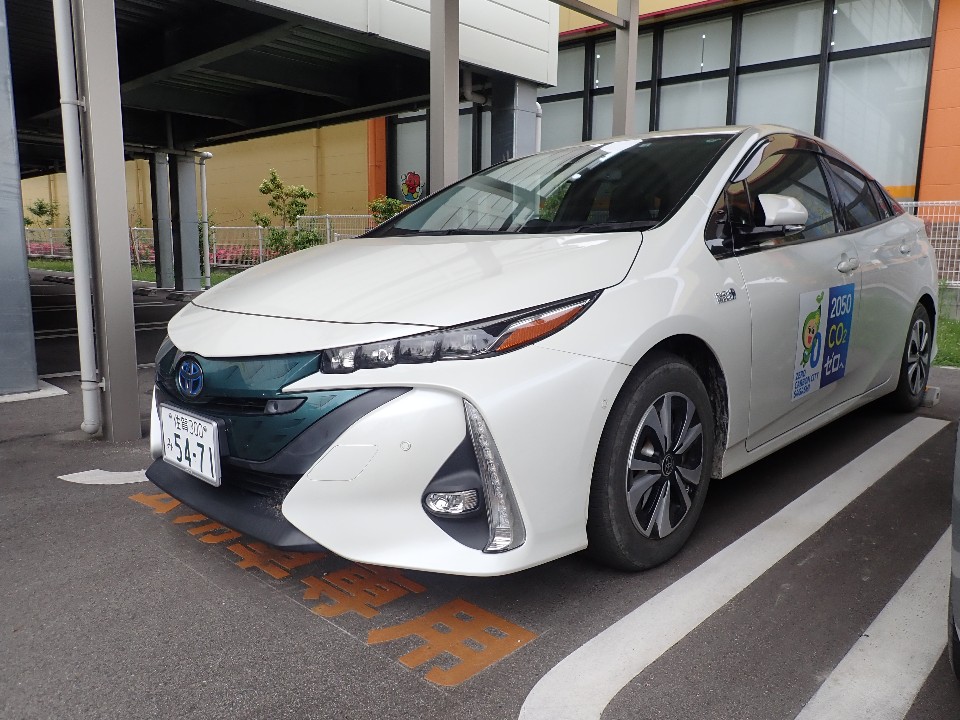 白いトヨタプリウスPHVが建物入口の駐車スペースに停車し、ドア側面に2030CO2削減目標達成のステッカーが貼られいる写真
