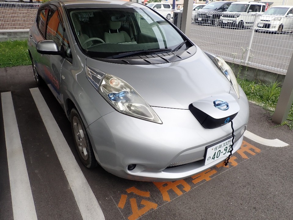 シルバーの日産e-NV200がオレンジ色の文字の駐車場に停車し、充電ケーブルが接続し充電中の写真