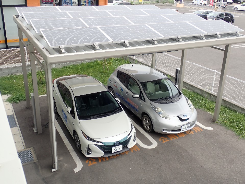 太陽光パネルを備えたカーポートの下に、白いトヨタプリウスPHVと日産リーフの2台が駐車スペースに停車している電気自動車充電ステーションの写真