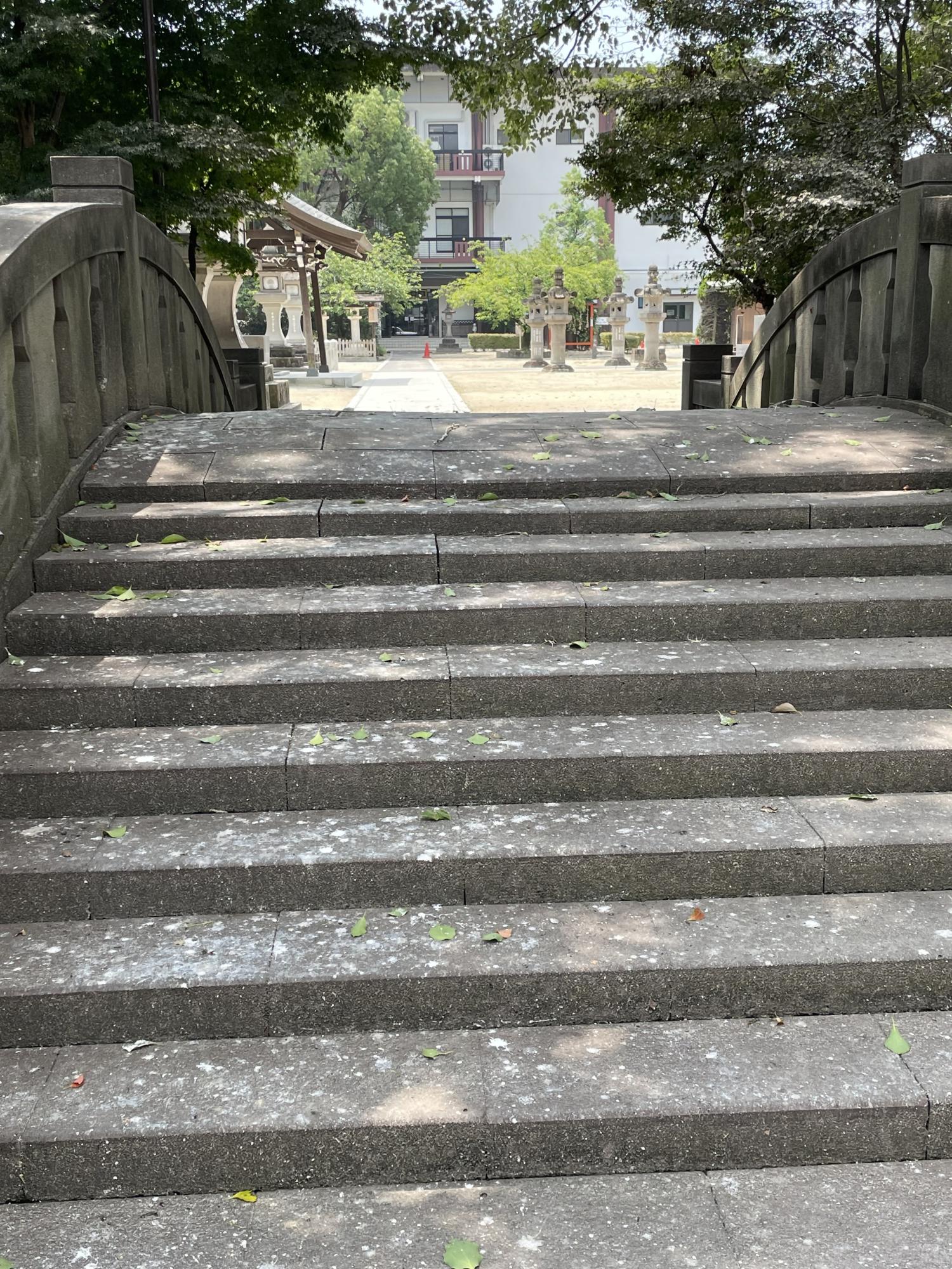 石灯籠が並ぶ広場や伝統的な建築物、木々の緑が広がる神社の境内のような風景が奥行きを持って映し出されている手前の、石造りの太鼓橋の階段にサギの糞が広がって落ちている写真
