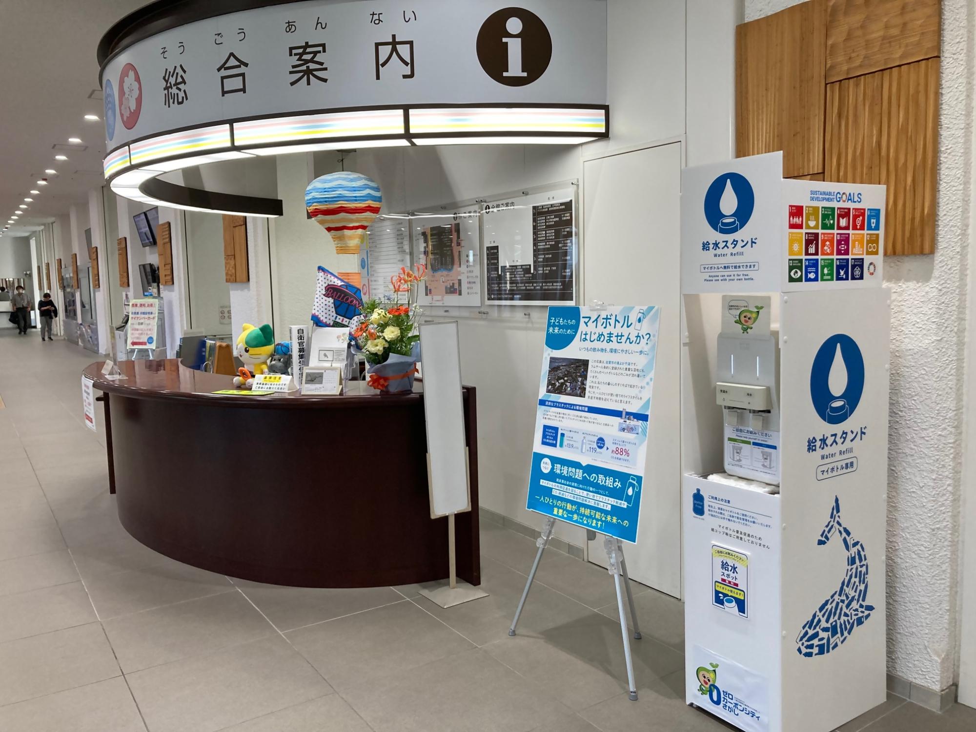 「総合案内」と書かれたカウンターの横に、「給水スタンド」と表示されたマイボトル専用の白いウォーターサーバーと、マイボトル利用を推奨する啓発用パネルが設置されているウォーターサーバーの設置場所を示した写真