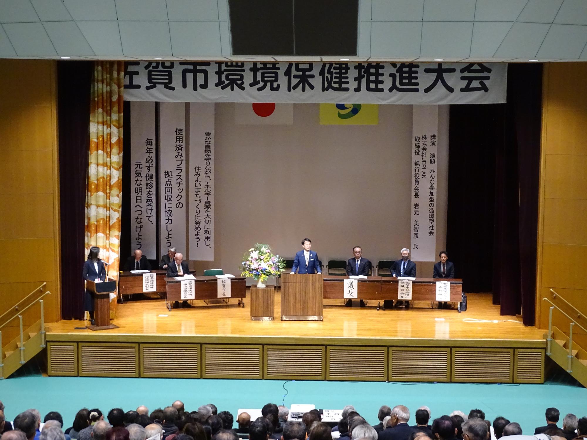 「佐賀市環境保健推進大会」と書かれた横断幕が掲げられた舞台上で、中央の演台に立つ男性がスピーチを行い、その左右には役職札が置かれた席に座る複数の来賓と、客席を埋め尽くす多くの参加者の様子を捉えた大会の様子の写真