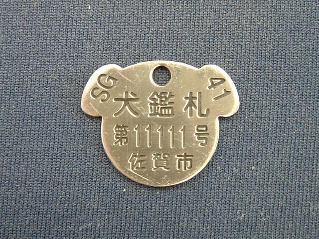 犬の顔型をした金色の金属製鑑札に犬鑑札の文字と第11111号および佐賀市の刻印が施された犬の登録証の写真