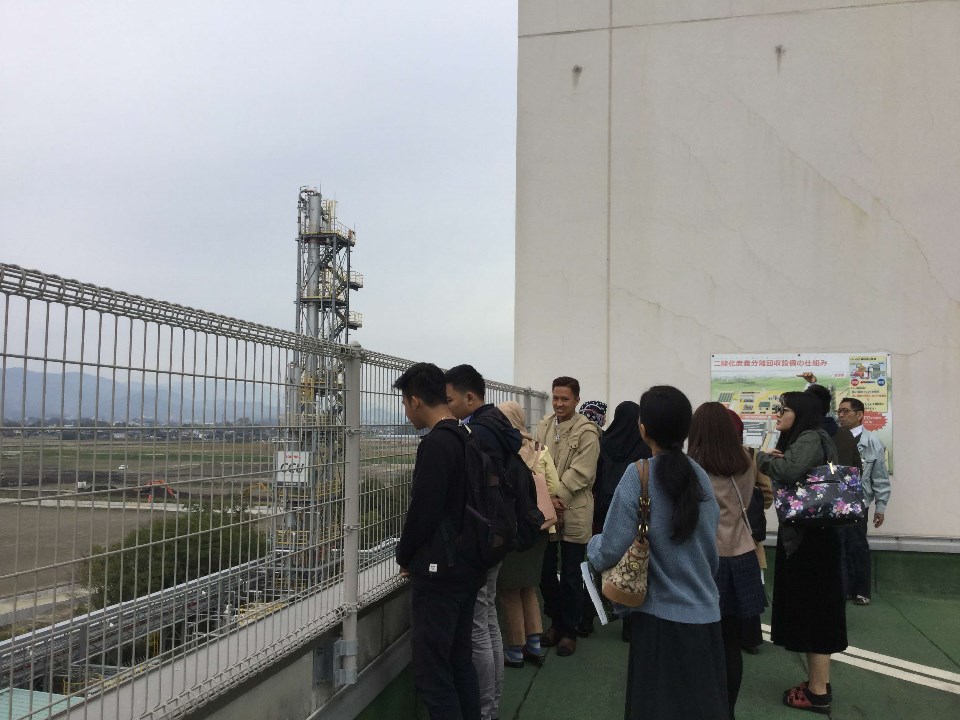フェンス越しに高い塔のようなプラント設備が立つ広大な敷地を、リュックを背負った若者やカバンを持った女性など、多様な服装の人々が建物の屋上から見学している、施設見学会の様子を捉えた写真