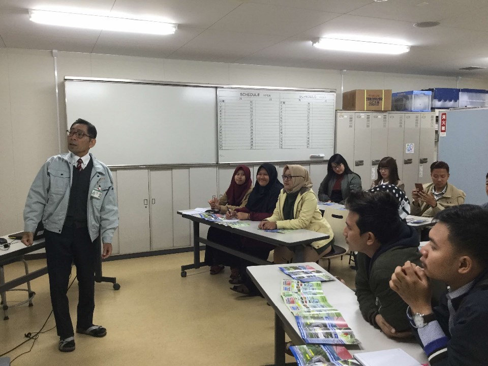 作業着を着た男性がホワイトボードの前で説明を行い、机に並んだパンフレットを手に取りながら、ヒジャブを被った女性を含む多国籍な受講生たちが熱心に話を聞いている、室内での研修や説明会の風景を捉えた写真
