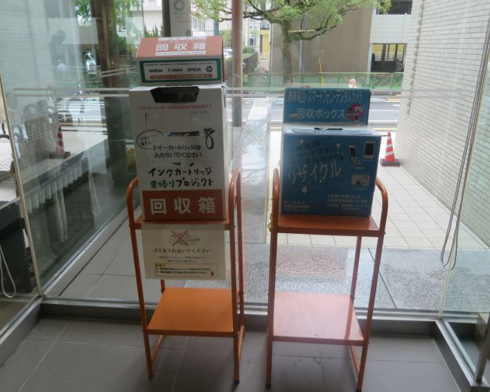 建物のガラス窓際に設置された、左側にインクカートリッジ里帰りプロジェクトの白い回収箱、右側に携帯電話やスマートフォン、デジタルカメラを回収するための青いリサイクルボックスが、それぞれオレンジ色の専用什器に載せて並べられている写真