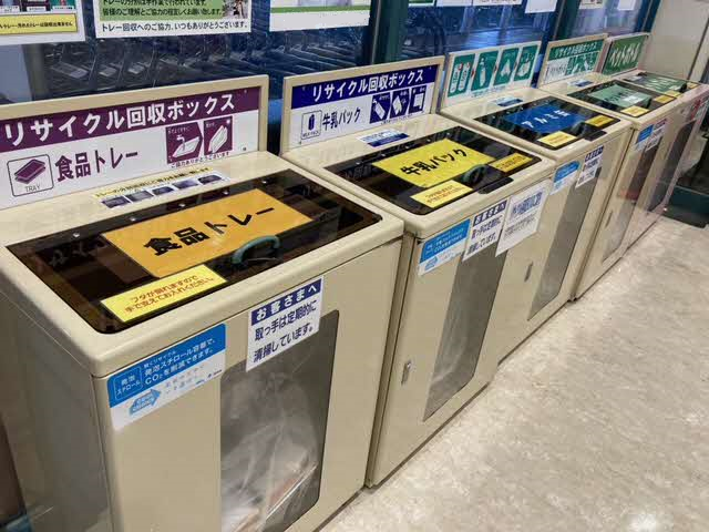 店内に「食品トレー」「牛乳パック」「アルミ缶」「ペットボトル」と分別ラベルが貼られたリサイクル回収ボックスが横一列に並び、中身が見える透明な窓がついたベージュ色の専用容器が設置されている写真
