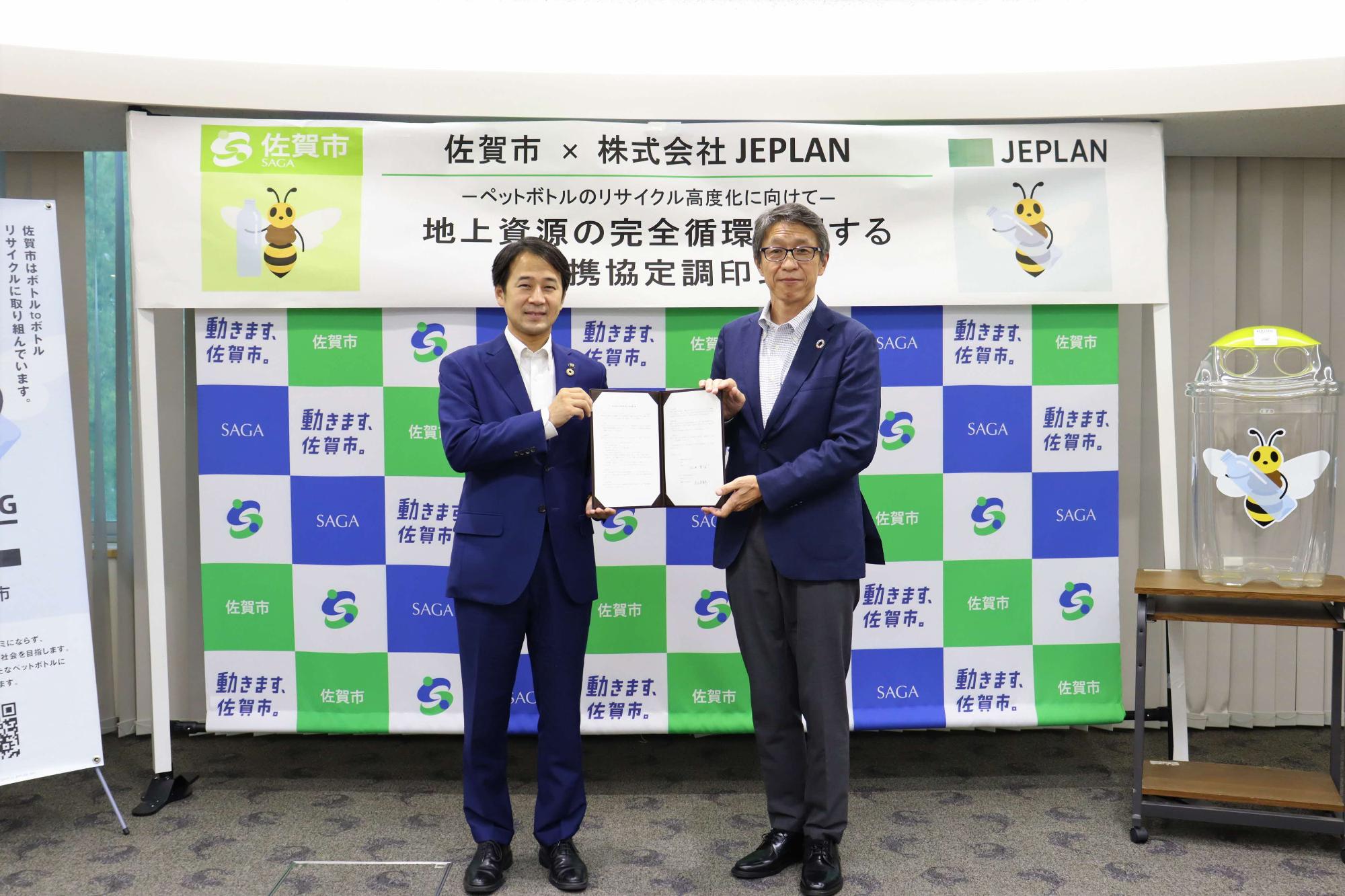 佐賀市のロゴと株式会社JEPLANのロゴが交互に配置されたバックボードの前で、スーツ姿の市長と岩元美智彦様が連携協定の署名文書を手に並んで立ち、ペットボトルの完全循環に向けた協力体制をアピールしている記念写真