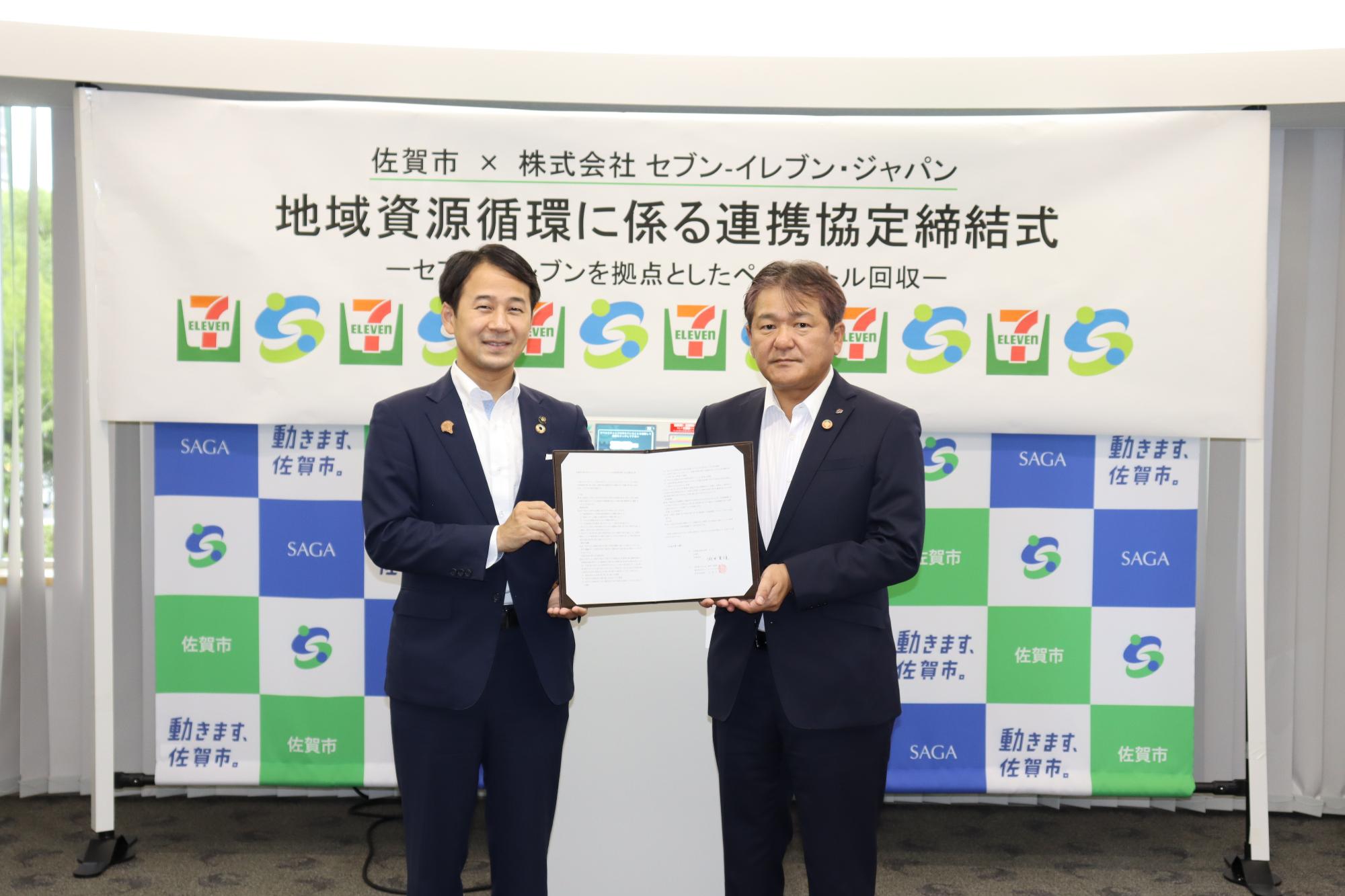 佐賀市と株式会社セブン-イレブン・ジャパンによる地域資源循環に係る連携協定締結式にて、スーツ姿の市長と桝尾 威彦様が署名された協定書を手に持ち、ロゴが並ぶ横断幕と市松模様のバックボードの前に並んで記念撮影をしている写真
