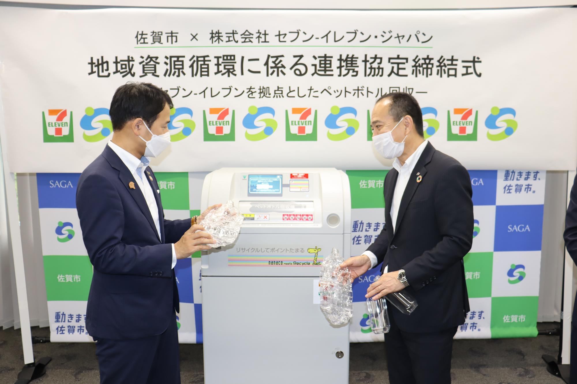 佐賀市とセブン-イレブンによる地域資源循環の連携協定締結式にて、スーツ姿の男性二人がペットボトル自動回収機を挟んで立ち、リサイクル用のペットボトルを手に持ちながら機械の動作を確認している様子の写真