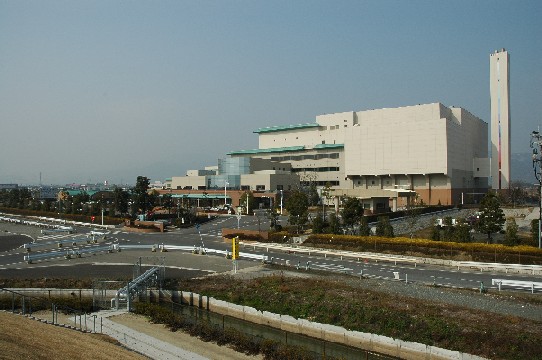 平坦な土地に建つ、淡いベージュの外壁と緑色の屋根が特徴的な多層構造の巨大な工場建築が映し出されており、右端には四角柱の細長い煙突がそびえ立ち、手前には舗装された道路や整理された植栽が広がる、佐賀市清掃工場の外観写真