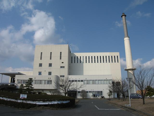 白を基調とした大規模な清掃工場の建物と高く伸びた煙突が映し出されており、手前には植栽が整えられたアプローチや数本の枯れ木が並ぶ広々とした敷地が広がっている佐賀市清掃工場南部中継所の外観写真