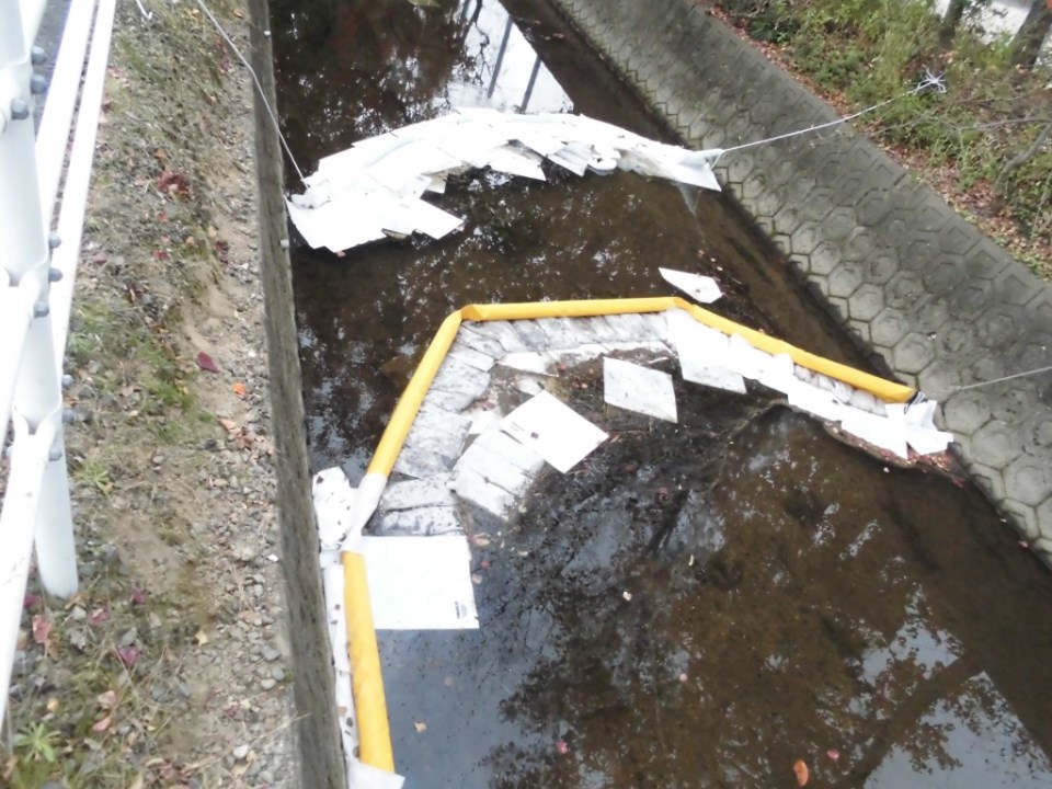 水路にて、流出した油の拡散を防ぐために黄色いオイルフェンスが張られ、その内側や上流側に白い吸着マットが多数浮かべられて水面を覆っている、油回収作業中の現場を捉えた写真