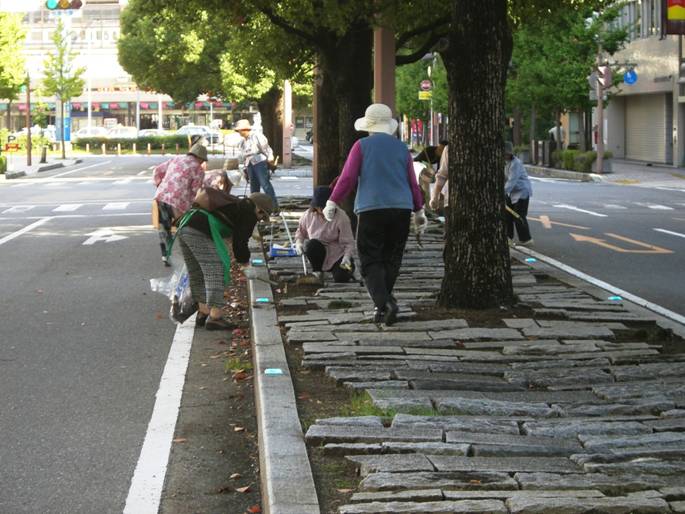市街地の道路沿いで、複数の市民たちが、等間隔に木が植えられた石畳の歩道にしゃがみ込み、協力して熱心に清掃活動や草取りを行っている様子の写真