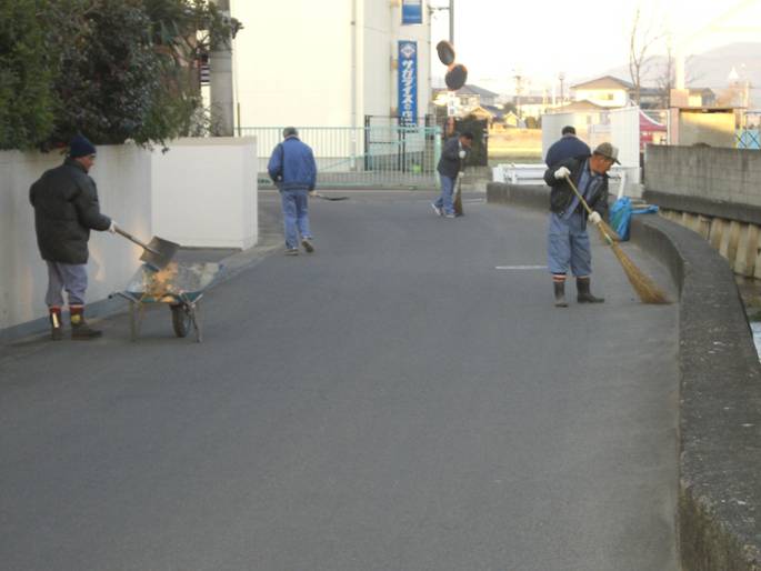 住宅街の道路で、数人のボランティアが竹ぼうきで路面を掃いたり、シャベルを使って集めたごみや落ち葉を一輪車へ積み込んだりして、美化活動に取り組んでいる様子を捉えた写真