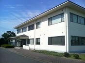 白く角ばった2階建ての衛生センター管理棟が青空の下にあり、その手前には舗装路と低木の植え込みが配置されている写真