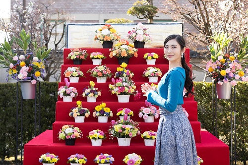 赤い布が敷かれた雛壇のような階段状の台に色とりどりの華やかなフラワーアレンジメントが多数並べられ、その横で青いトップスを着た女性が微笑みながらポーズをとっている写真