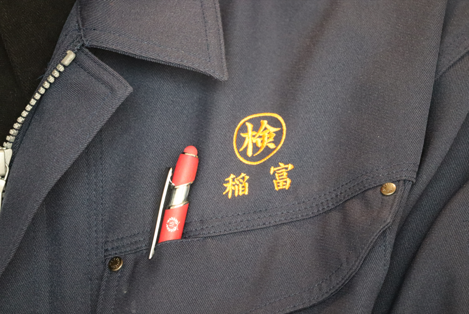 紺色の作業服の胸ポケットに、黄色い糸で丸囲みの「検」という文字と「稲富」という苗字が刺繍されており、ポケットには赤いペンが一本差し込まれている様子を至近距離から撮影した写真