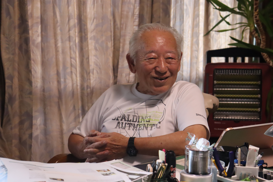 白いTシャツを着た白髪の篠原 清彦さんがデスクに座り、穏やかな笑みを見せている写真
