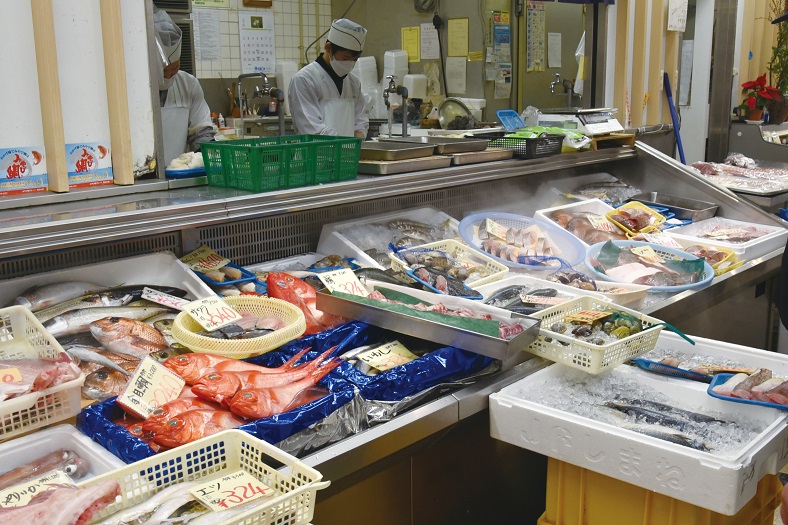 調理服と帽子を着用した店員が奥の調理場で作業を行い、手前のショーケースには氷を敷き詰めた容器に金目鯛や貝類などの多様な魚介類が所狭しと並べられている、清潔感のある鮮魚店の店内の写真