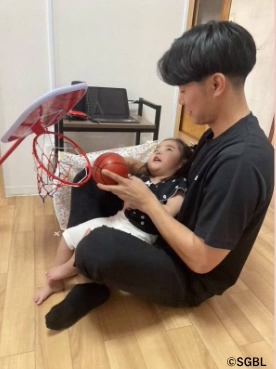 膝の上に抱えた幼い子どもと一緒に小さなおもちゃのバスケットゴールへ赤いボールを入れようと、優しく導きながら触れ合っている井上選手を写した写真