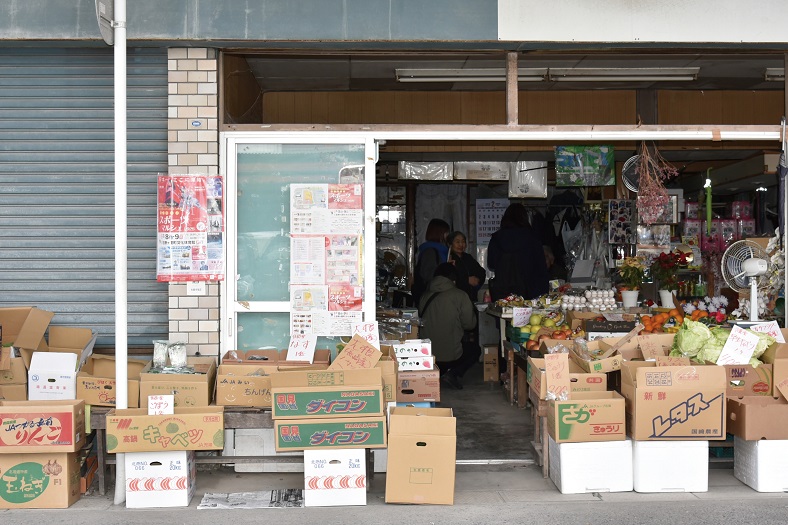 商店の店先に、ダイコンやレタスなどの野菜が入った段ボール箱が所狭しと並べられ、奥の薄暗い店内では数人の客が商品を品定めしている、活気ある昭和レトロな雰囲気漂う商店の店頭の写真