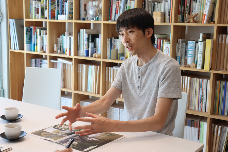 たくさんの本が並んだ本棚を背景にテーブルの席に座った川崎さんが身振り手振りを交えて熱心に話をしている写真