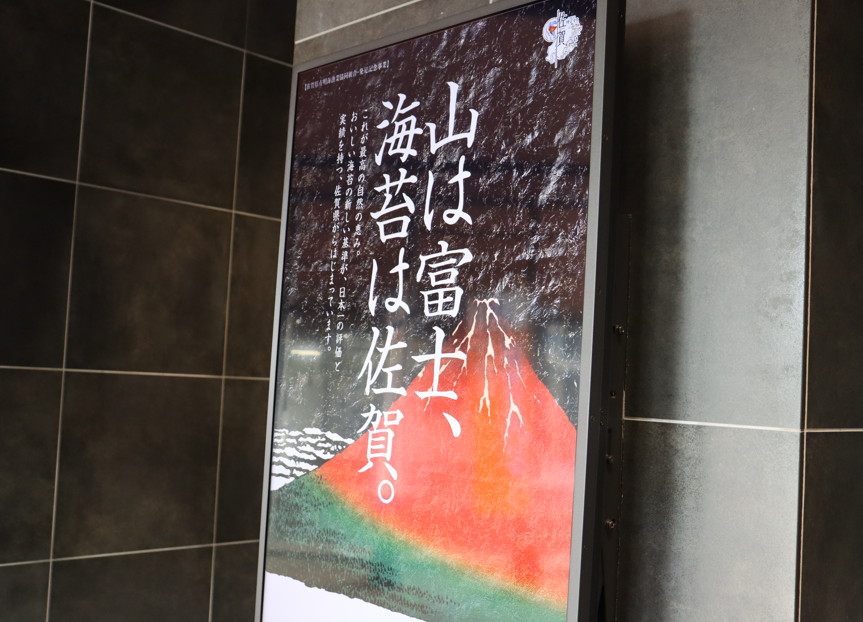 「山は富士、海苔は佐賀。」という大きな白い文字のキャッチコピーと赤富士のイラストが描かれた、広告ポスターを斜めから捉えた写真