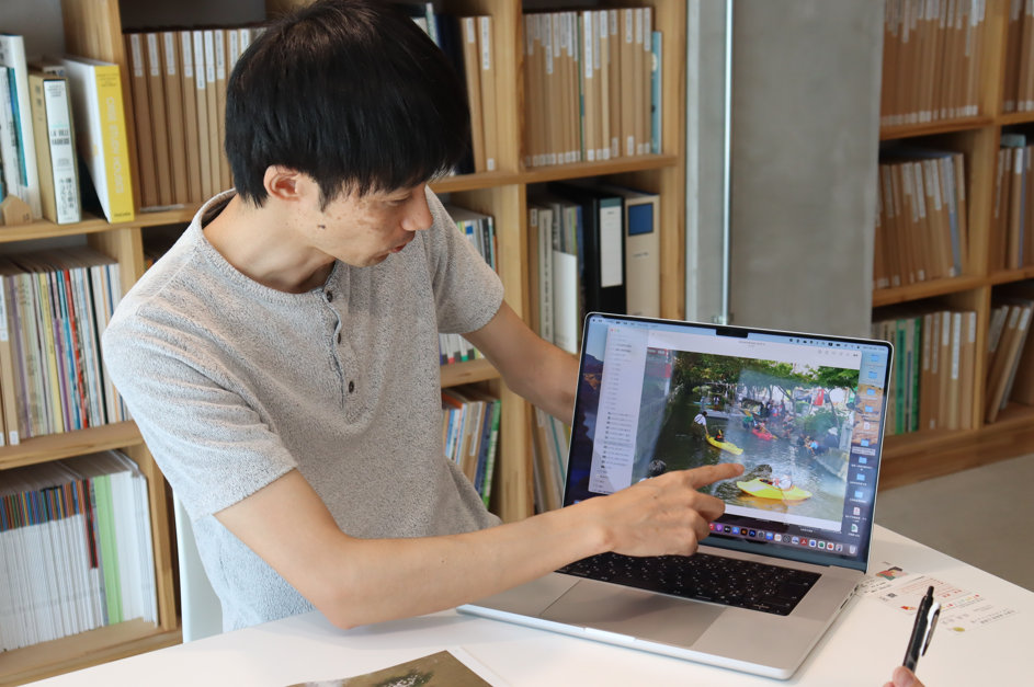 ノートパソコンの画面に表示された「水路でカヤックを楽しむ人々」の写真を指さしながら内容を説明している川崎さんの写真