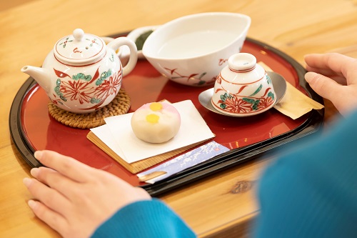 木製テーブルの上で、赤いお盆に載せられた花柄の急須と茶器、白い和菓子、そして湯冷ましがセットされた伝統的なお茶の膳を、青い服を着た人物が両手を添えて今まさに楽しもうとしている様子の写真