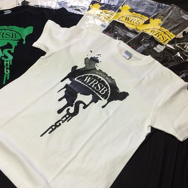 白と黒のTシャツが広げられ、中央の白いTシャツには佐賀県の形状を模した影に「WRSB」のロゴが重なるグラフィックが大きくプリントされており、背後には黄色いラベルの付いた袋入りの商品が複数並ぶ、三重津みやげホスモスカモスの商品の写真