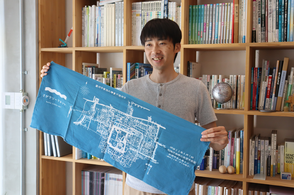 たくさんの本が並んだ本棚を背景に、「さがクリークネット」のロゴや佐賀市街図が白くプリントされた青い手ぬぐいを両手で広げて持ち、笑顔を見せる川崎康広さんの写真