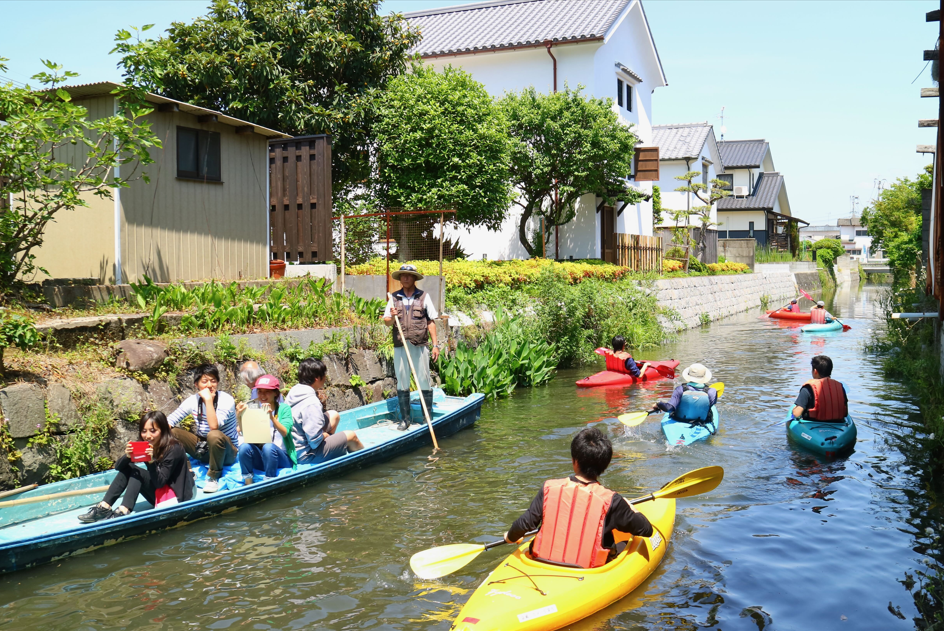 住宅が並ぶ水路で、カヤックに乗った人たちと、複数人が乗るボートが行き交い、水辺での活動を楽しむ様子を写した写真