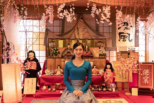 背景には屏風や提灯、掛け軸などが配置されたひなまつりの豪華な雛人形や桃の花が飾られた和室で、青い服を来た女性が緋毛氈の前に正座して微笑んでいる写真
