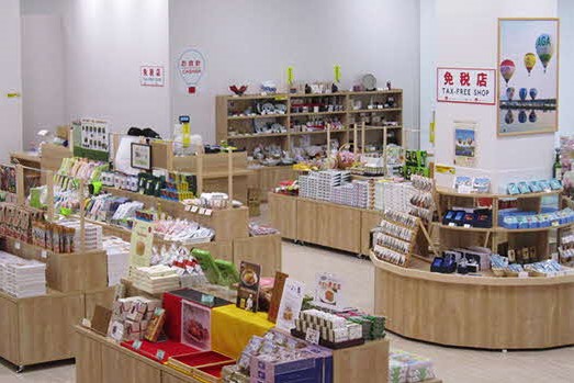 木の温もりを感じるベージュの棚に佐賀の特産品が整然と並び、免税店の看板やバルーンの写真が飾られた白い柱を中心に、整然とした陳列棚が広がる明るく清潔感のある佐賀工房店内の写真