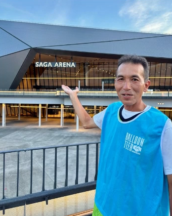 「SAGA ARENA」のロゴが輝く近代的なアリーナを背景に、水色の「BALLOONTEER」ビブスを着用した藤木さんが、建物を右手で指し示しながら紹介している様子を写した写真