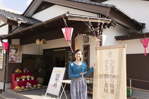 瓦屋根の和風建築の前で、青い服と柄物のスカートを身に着けた女性が「ひなまつり特選おもてなし茶屋」と書かれた看板の隣に立ち、笑顔でこちらを向いて施設を紹介している写真