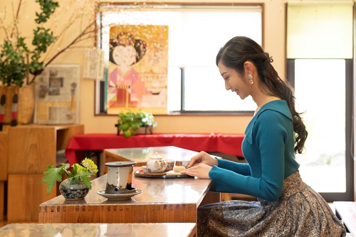 明るい店内の木製テーブル席で、青いトップスを着用した女性が横を向き、目の前に置かれたお盆の上のお茶セットや和菓子を優しく見つめながら、ゆったりとしたティータイムを楽しんでいる様子の写真
