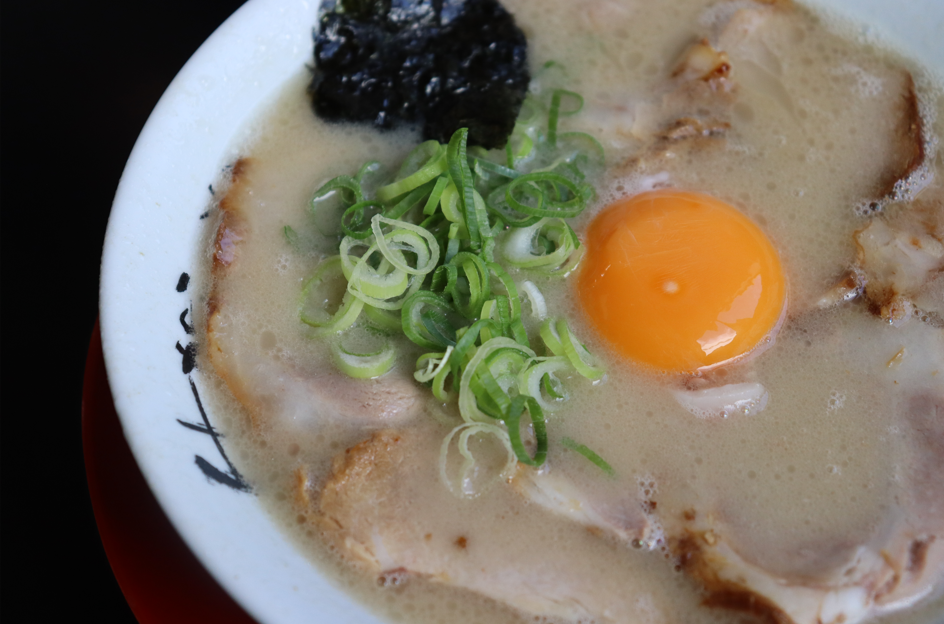白濁したとんこつスープと細切りのチャーシュー、鮮やかなオレンジ色の生卵の黄身、そしてたっぷりの刻みネギと海苔がトッピングされたラーメンを真上から写した写真