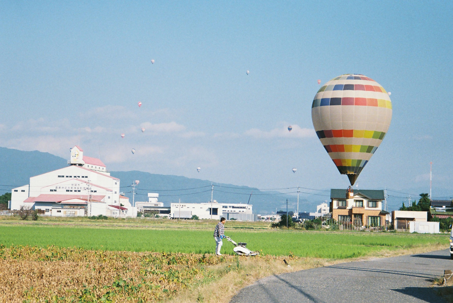 澄み渡る青空の下、のどかな田園地帯で農作業を行う人物の傍ら、カラフルなボーダー柄の大きな熱気球が低空を浮遊し、遠くの空にも多数のバルーンが点々と舞う風景を捉えた写真