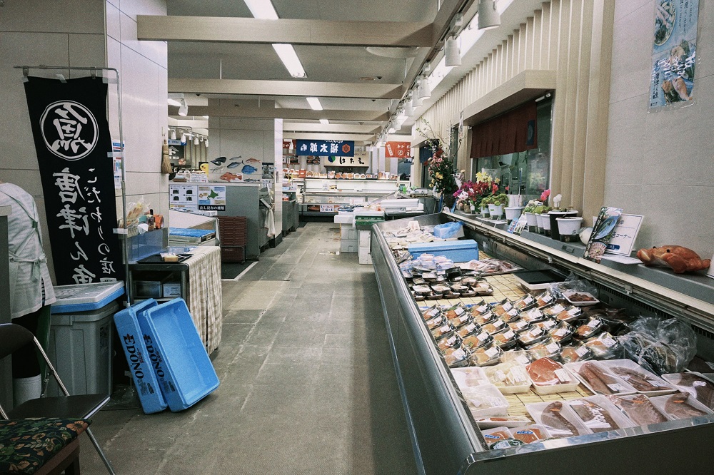 百貨店の鮮魚売り場にて、パック詰めされた切り身や干物が並ぶ長い冷蔵陳列ケースが奥へと続き、通路脇には黒い旗や青い運搬用ケースが立てかけられている清潔感のある店内の写真