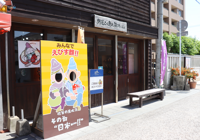 木造の「開運さが恵比寿ステーション」の建物前に、恵比寿様に扮した二人の人物の顔部分がくり抜かれた黄色い顔出しパネルが置かれており、その横には「ご自由にお入りください」と書かれた案内看板が立っている、観光スポットの様子を捉えた写真