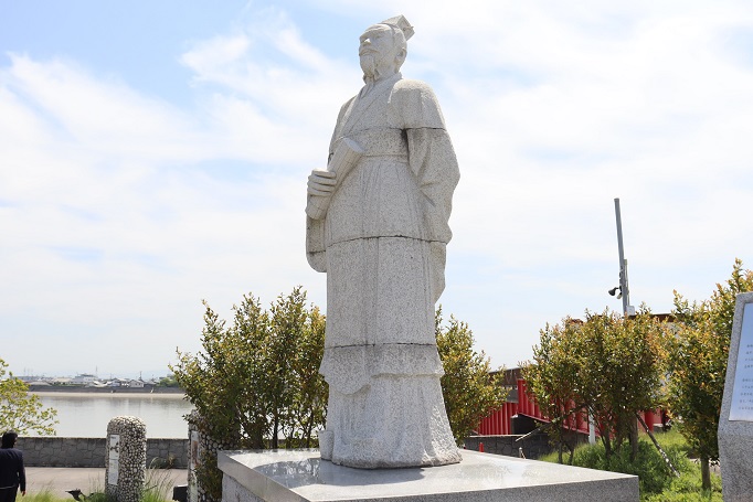 青い空と川を背景に、右手に巻物を持ち長い衣装を纏った古代中国の服飾スタイルの徐福の石像が、台座の上に立ち遠くを見つめるように設置されている、佐賀市諸富町にある徐福ゆかりの地の風景の写真